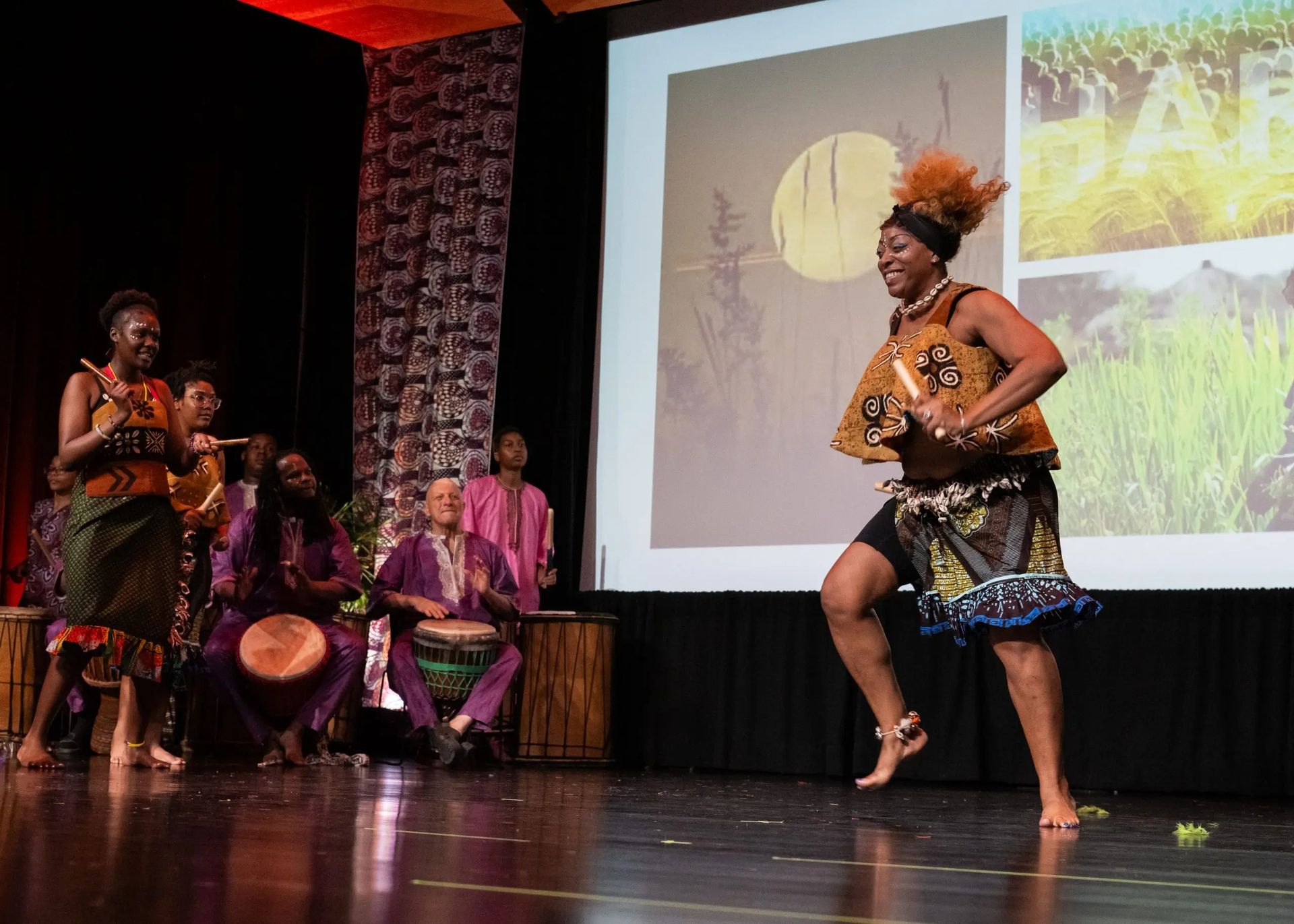 African dance performance: woman in costume dances on stage, musicians behind her, presentation screen background.