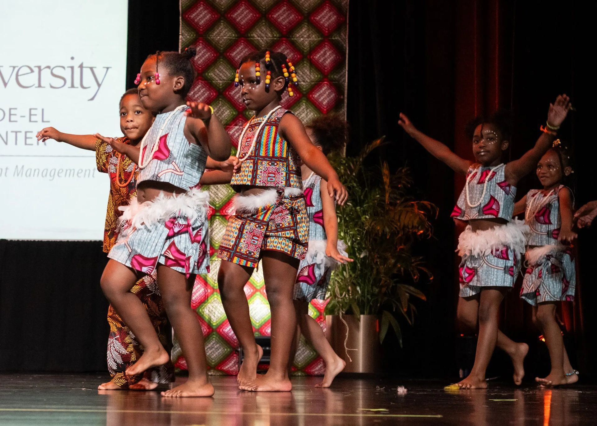 Children in colorful African-print costumes dance on a stage, arms raised, during a performance.