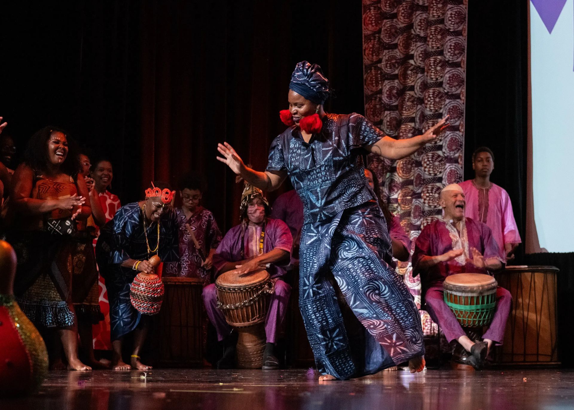 Woman dances on stage, surrounded by drummers. Vibrant costumes in purple, blue, and red.
