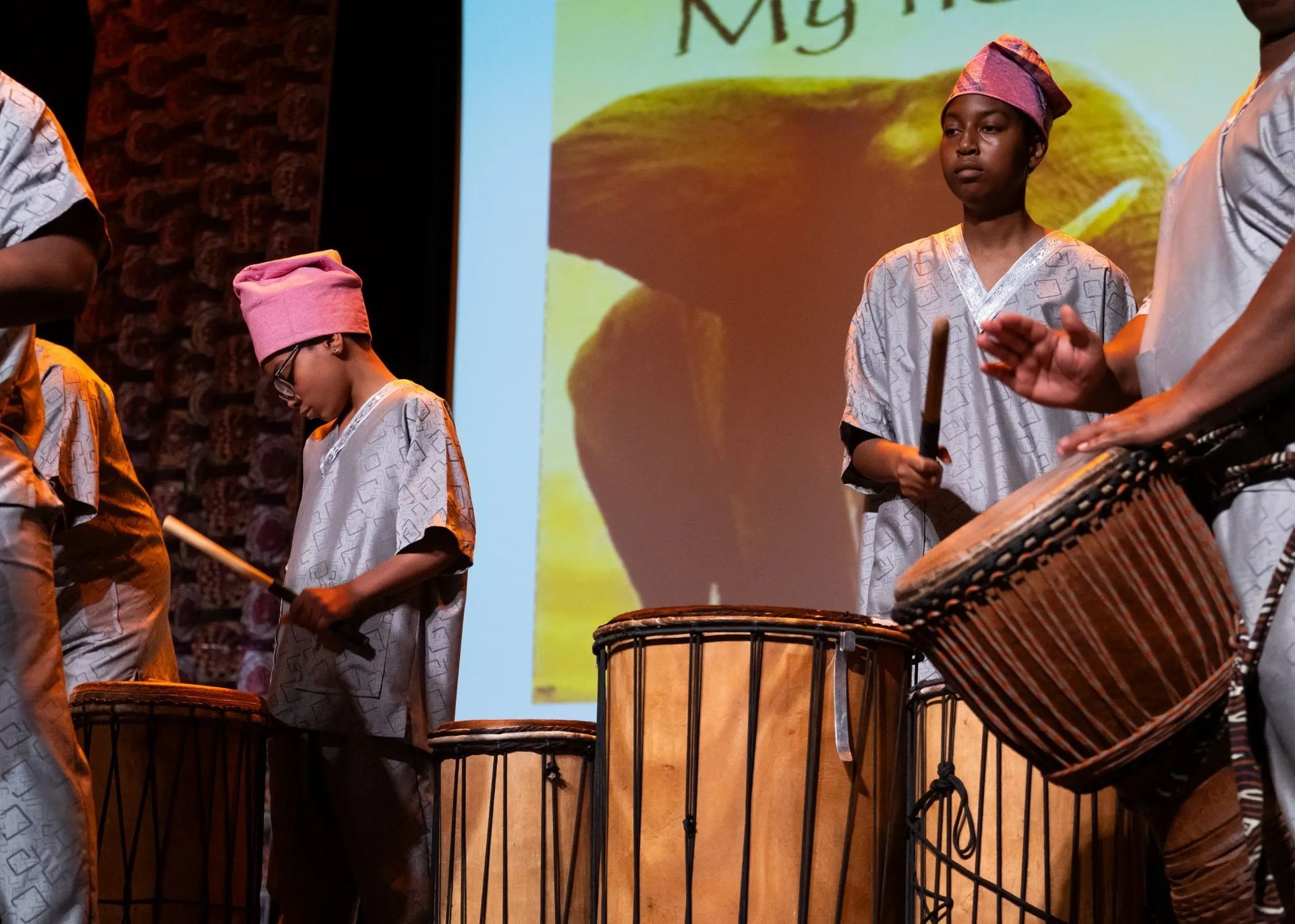 Group of Black people playing drums onstage; wearing matching silver outfits and pink hats.