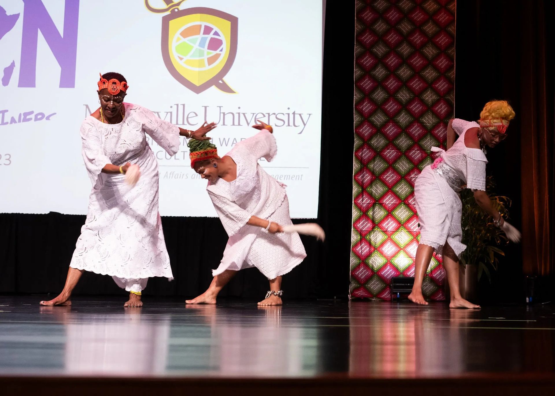Three women in white dresses perform a dance on stage; one holds flowers.