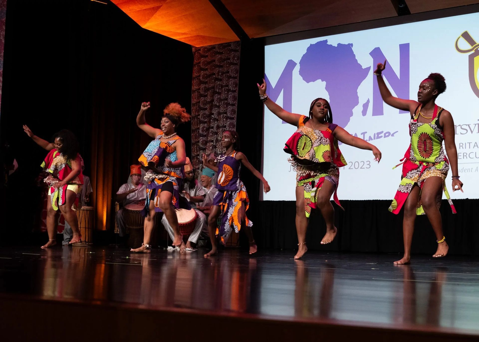 African dancers in vibrant outfits perform on a stage with musicians and a projected African map.