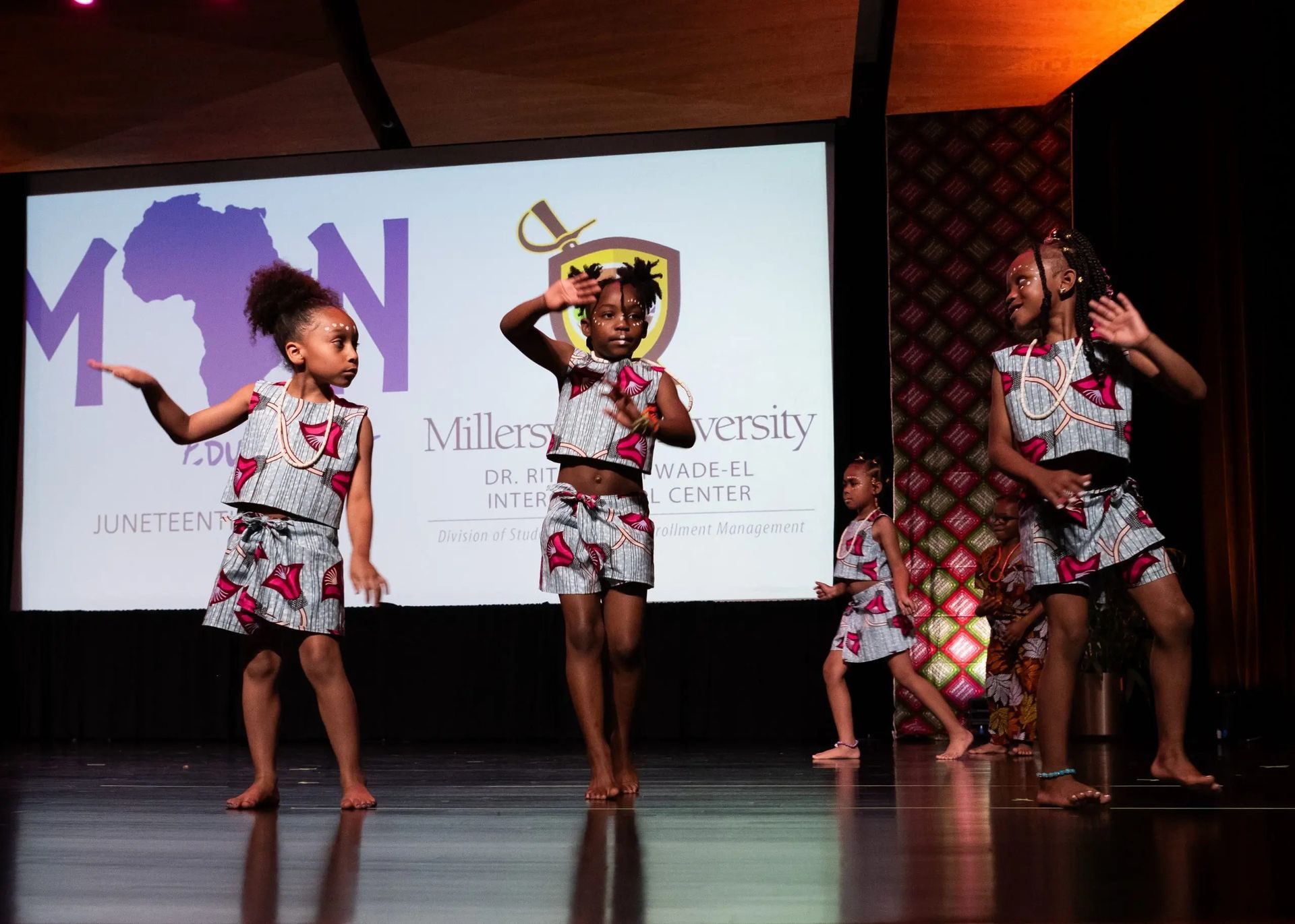 Young girls in matching patterned outfits dance on stage.