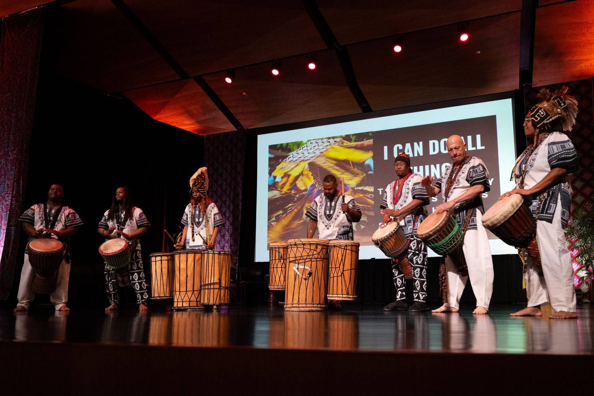 Group of people playing drums on a stage; banner reads 