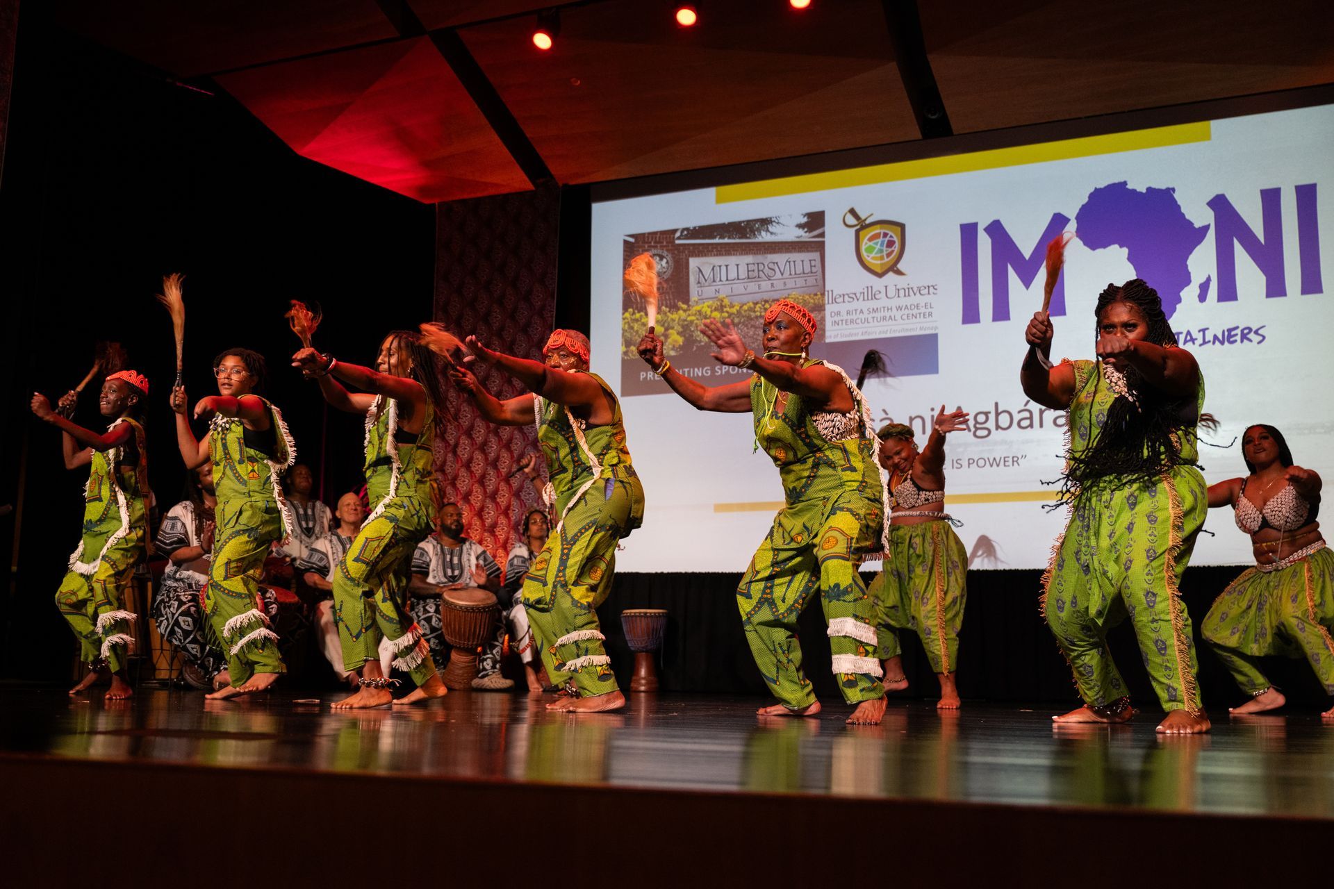 African dancers on stage wearing green patterned outfits, holding sticks, performing. A screen with a logo in the background.