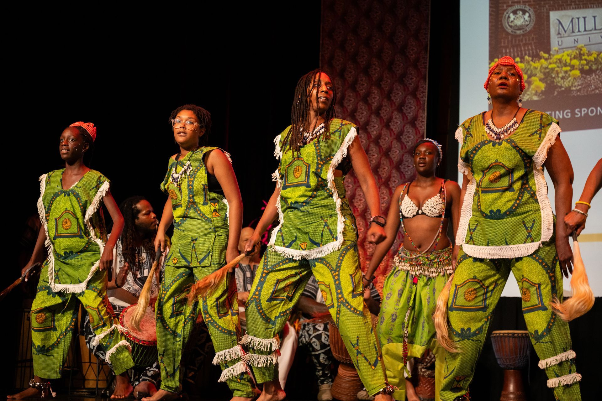 African dancers in green outfits perform on stage, with drummers in the background.