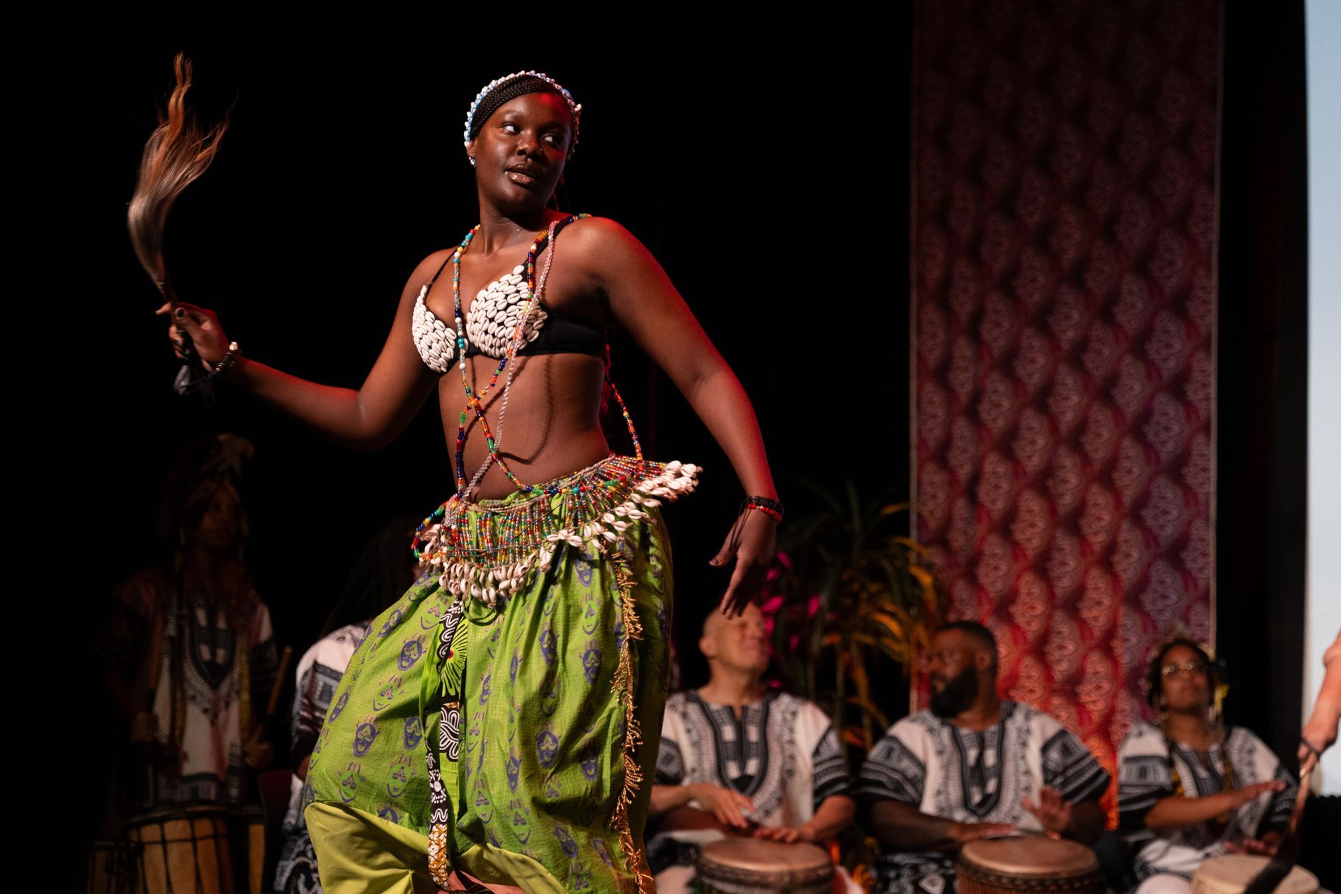 African dancer in green skirt and beaded top performs with drummers in background.