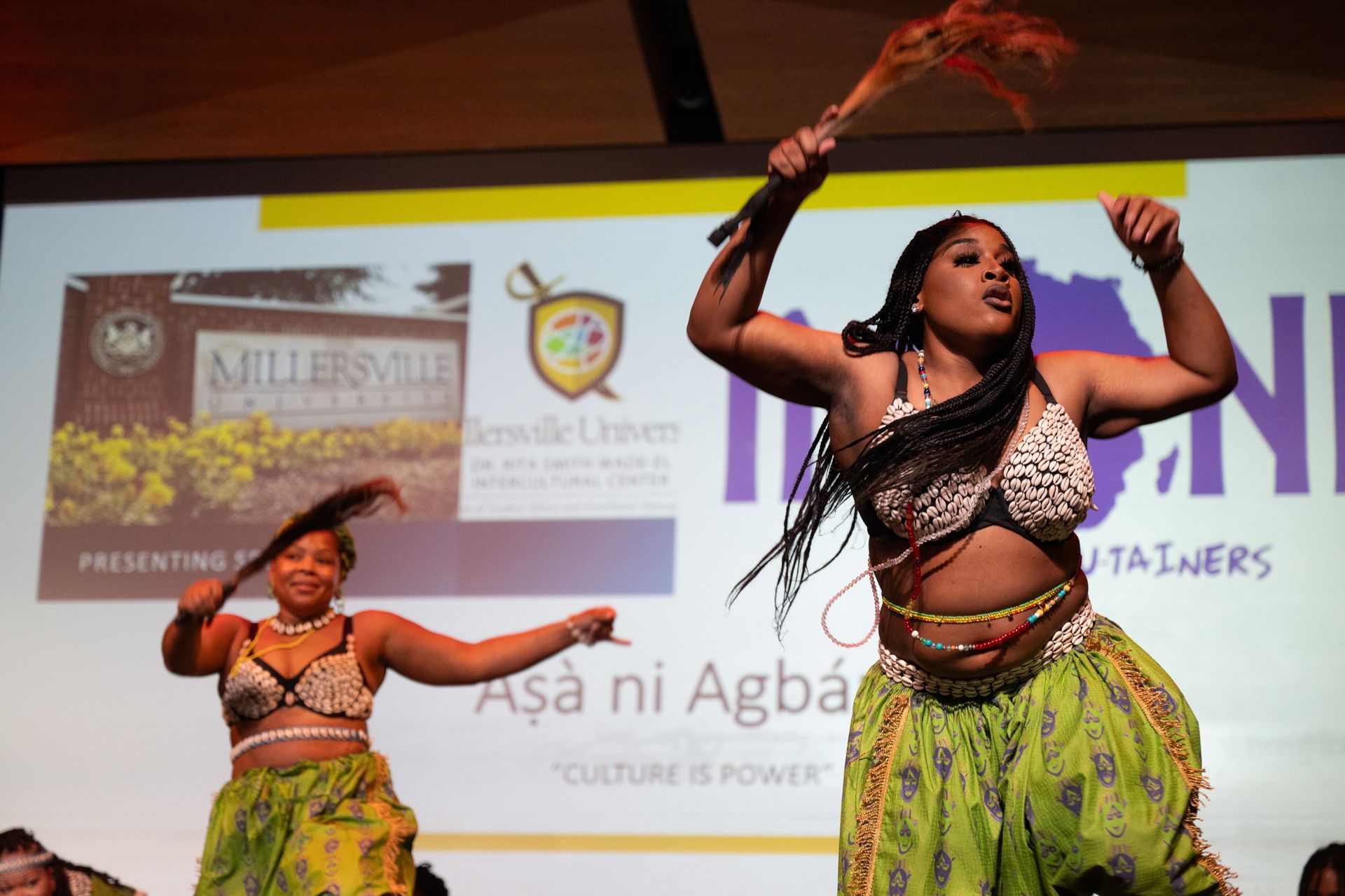 Two women perform a cultural dance on stage, wearing ornate outfits with bright colors and smiling.