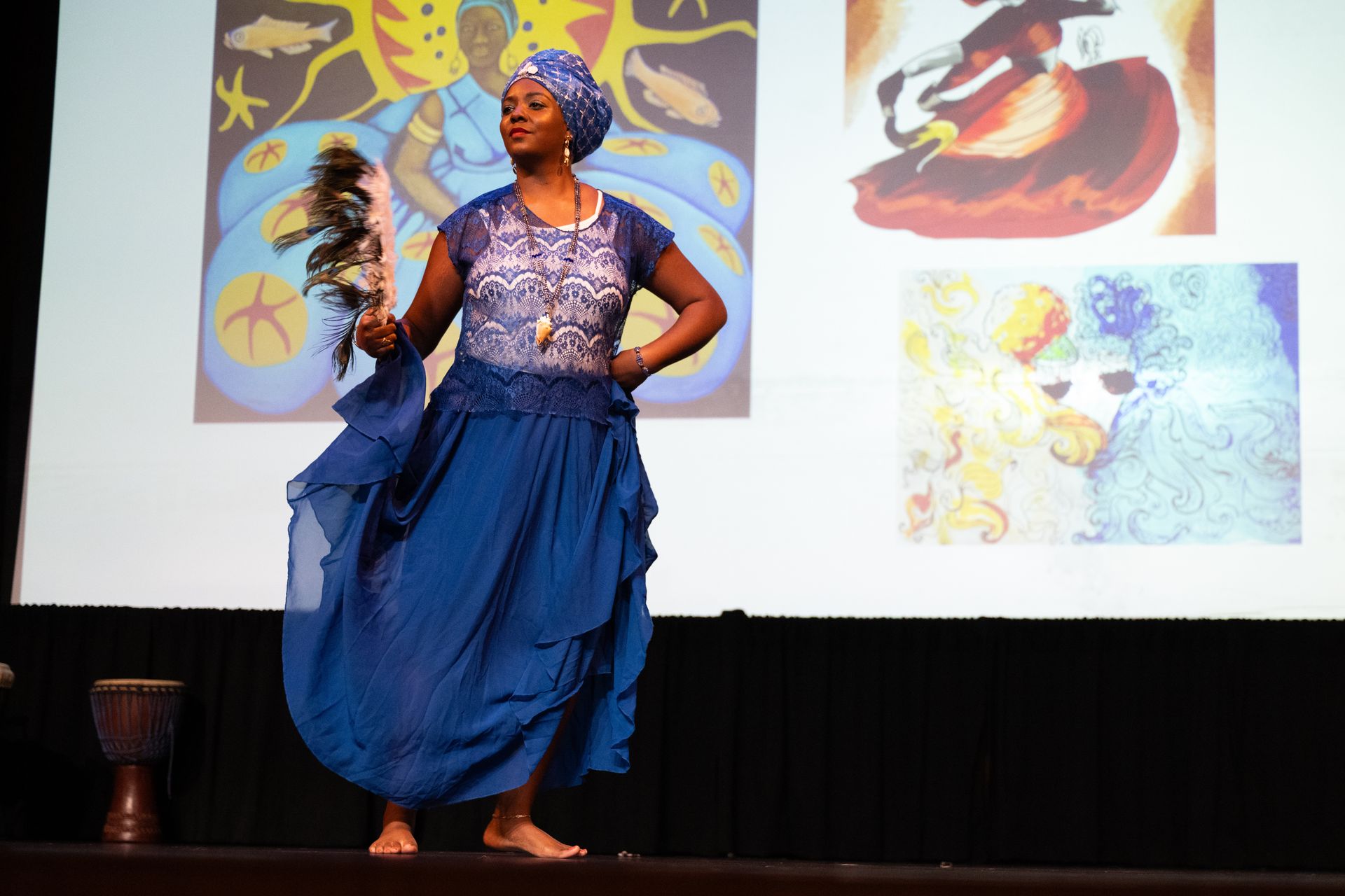 Woman in blue dress dances on stage, holding a fan, with artwork in the background.