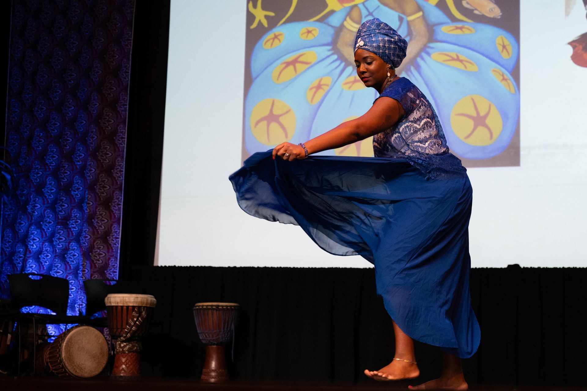 Woman in blue African attire dancing onstage, holding up her skirt; drums and art in background.
