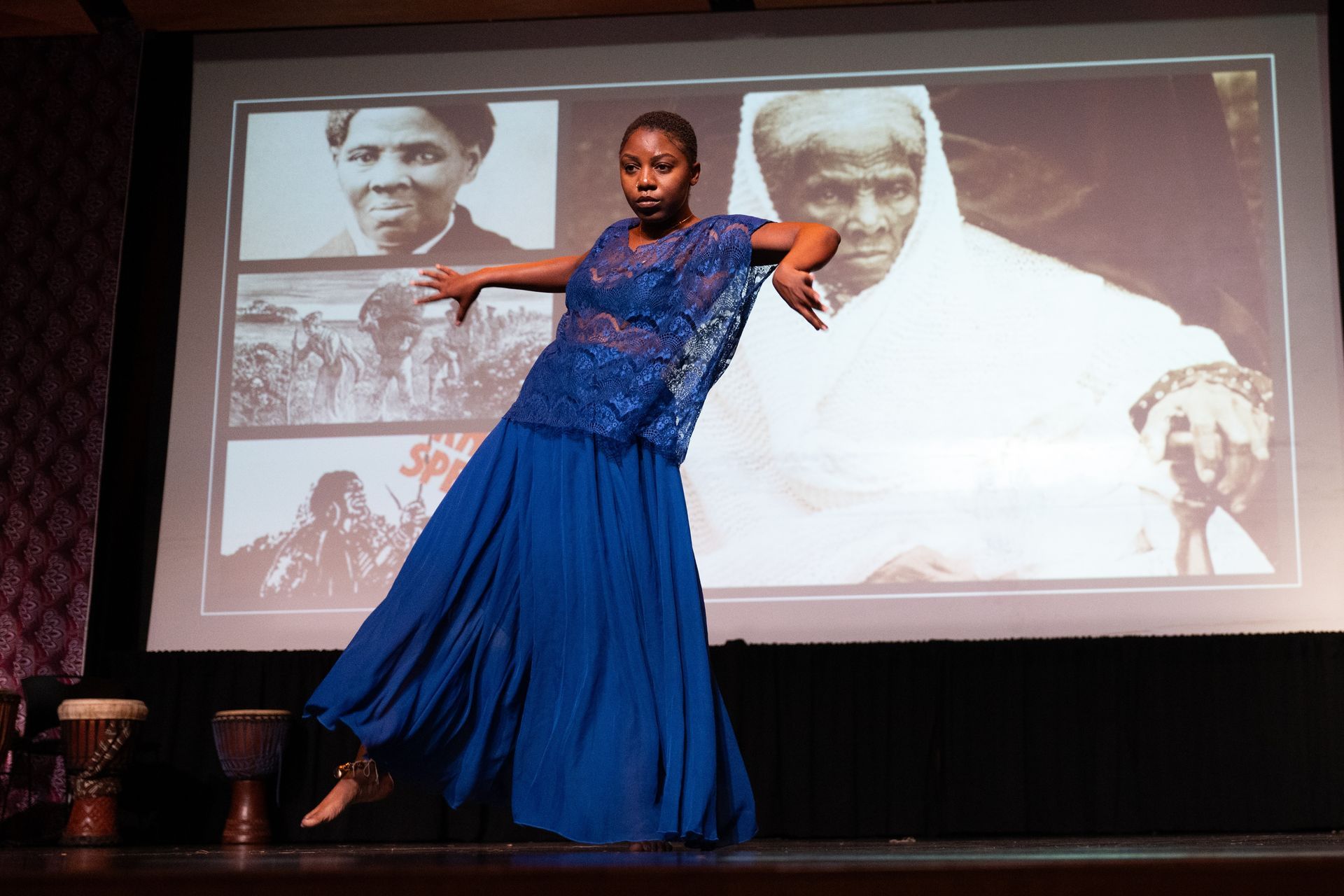A dancer in blue performs on stage with historical photos as a backdrop.