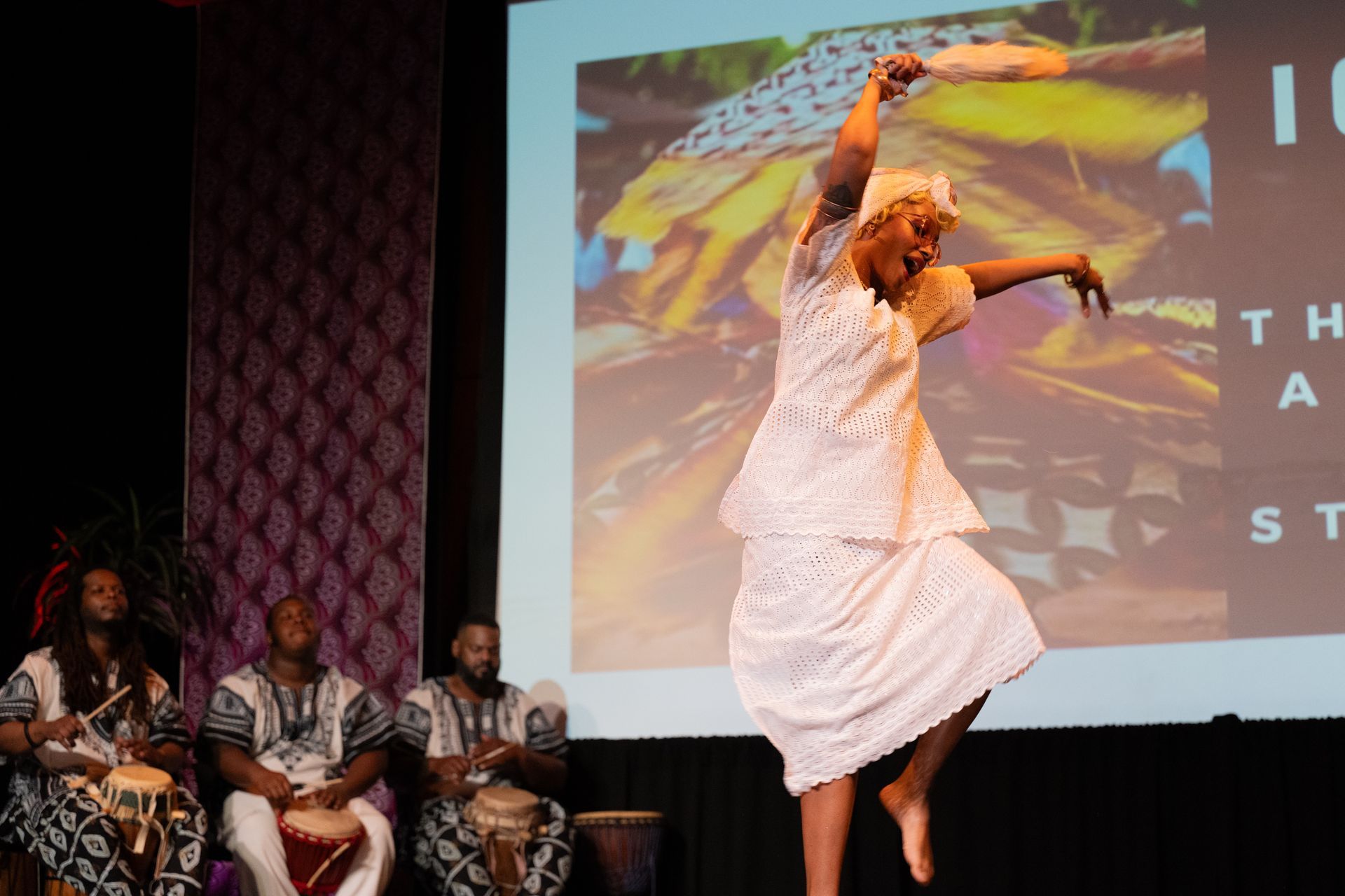 Woman in white dress dances with a fan as drummers perform on stage.