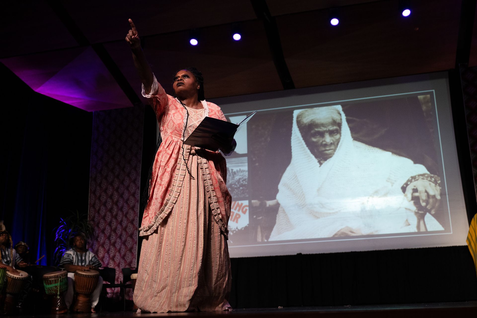 Woman in pink dress performs on stage with photo of elderly woman. Band and purple lighting.