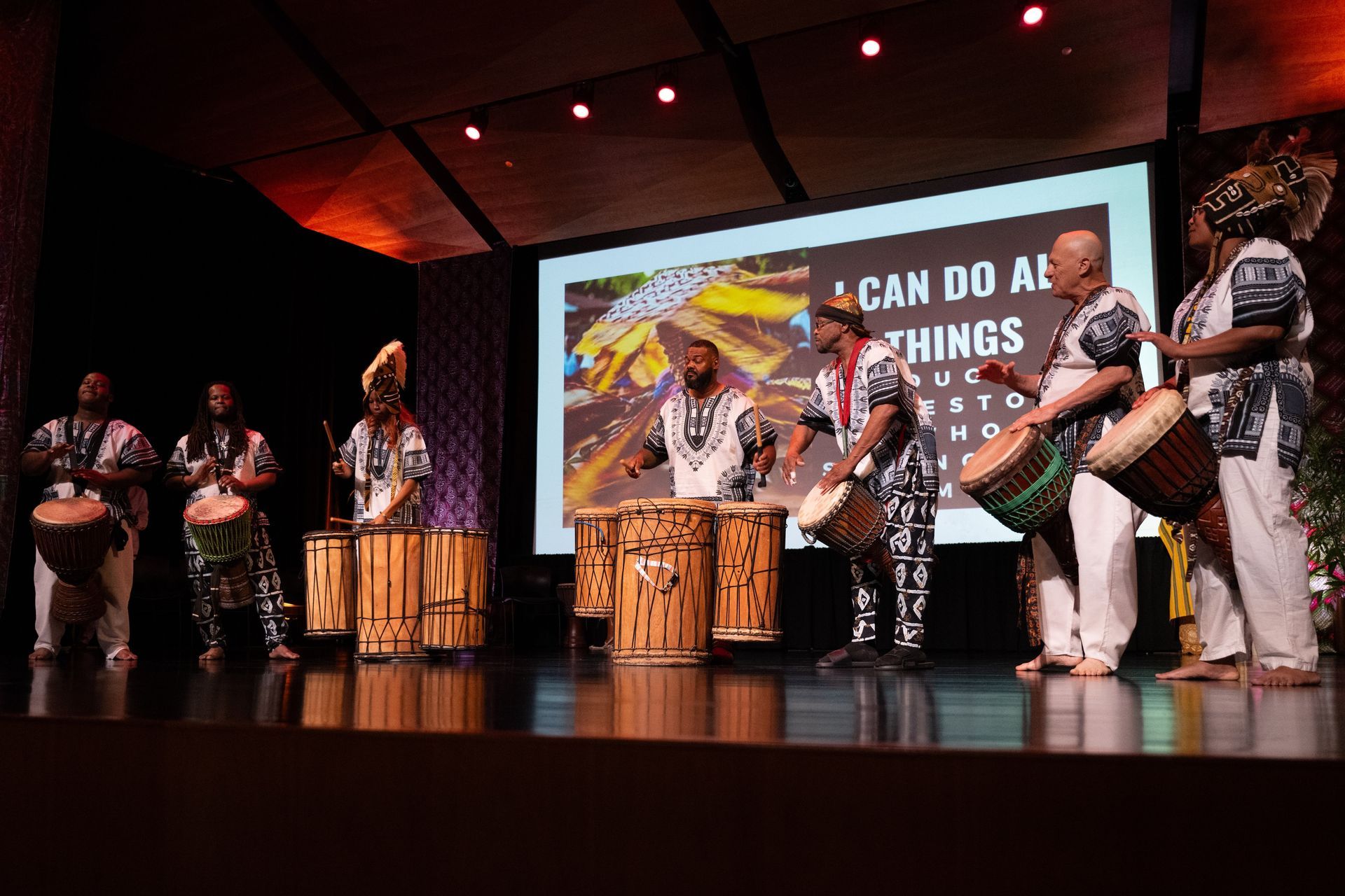 African drummers on stage, playing drums in a performance setting, with a screen behind them.