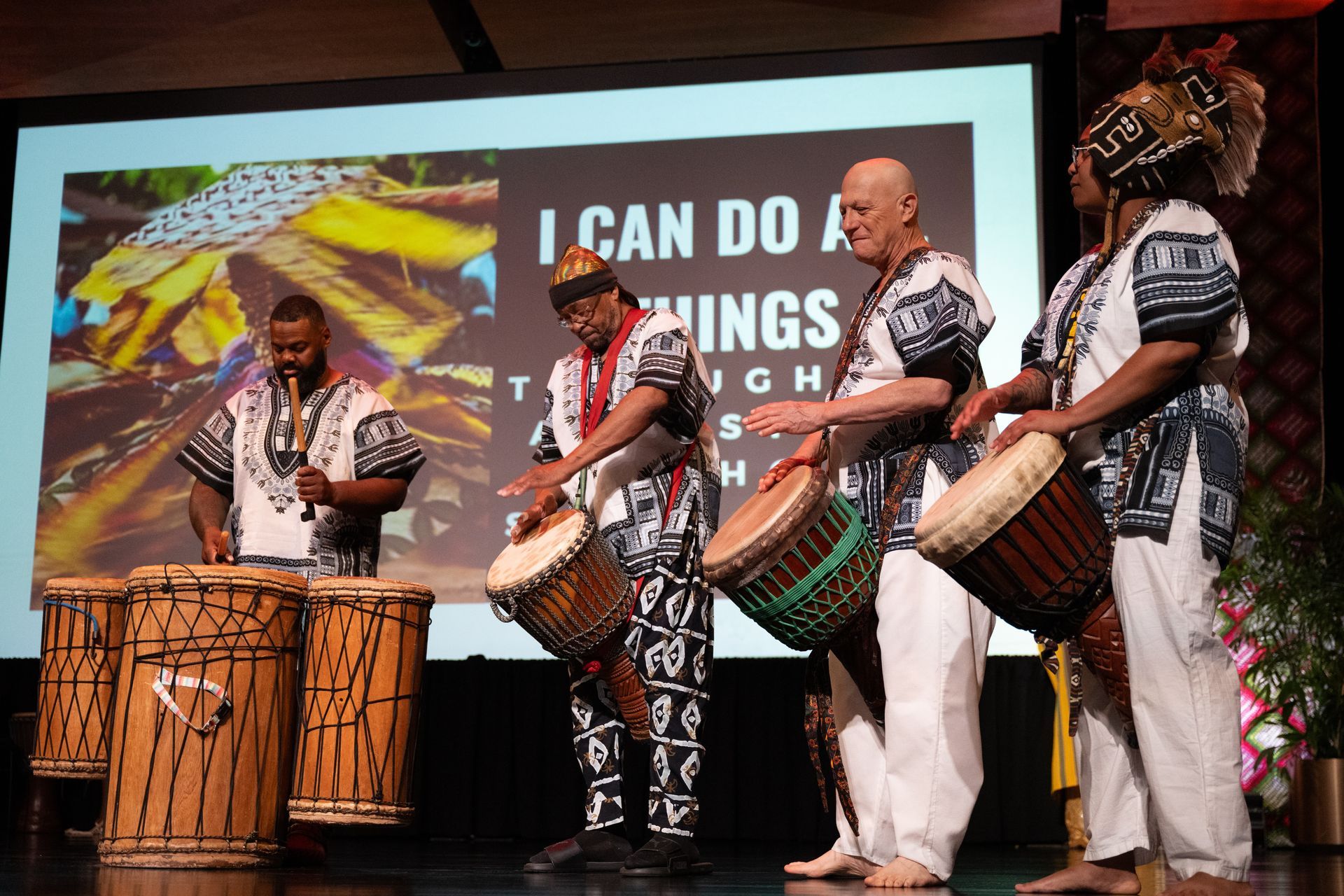 Four people in African attire drumming on stage; background shows a motivational message.