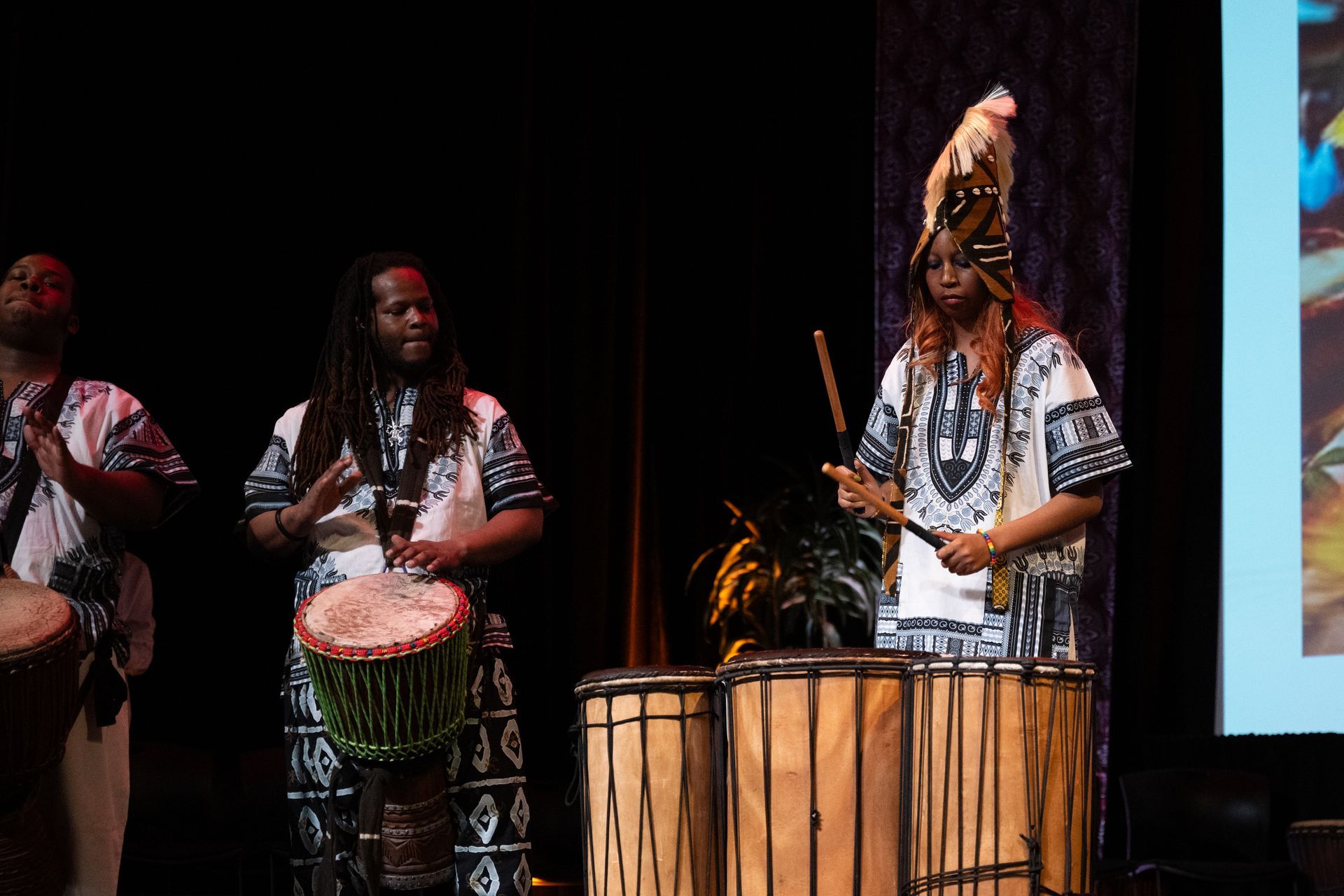 Three people in African attire playing drums on stage.