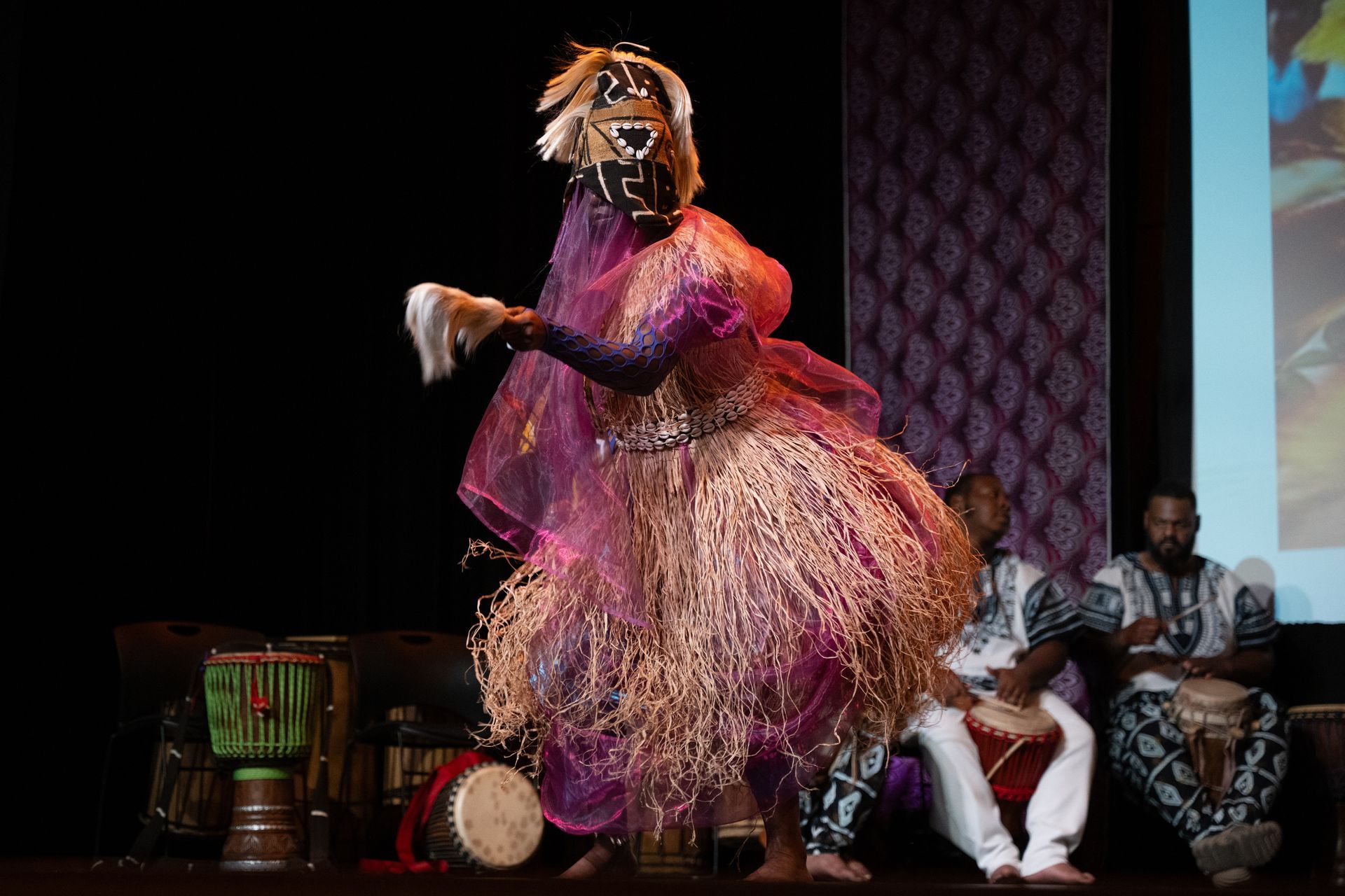 African dancer in purple and straw costume, masked, dancing on stage with drummers.