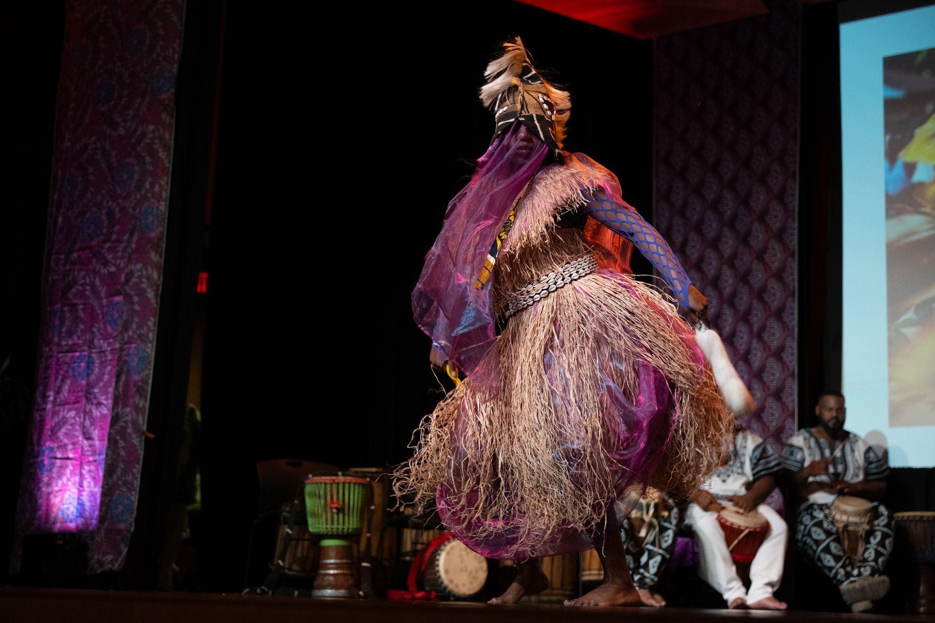 A costumed dancer performs on stage, surrounded by drums and musicians.