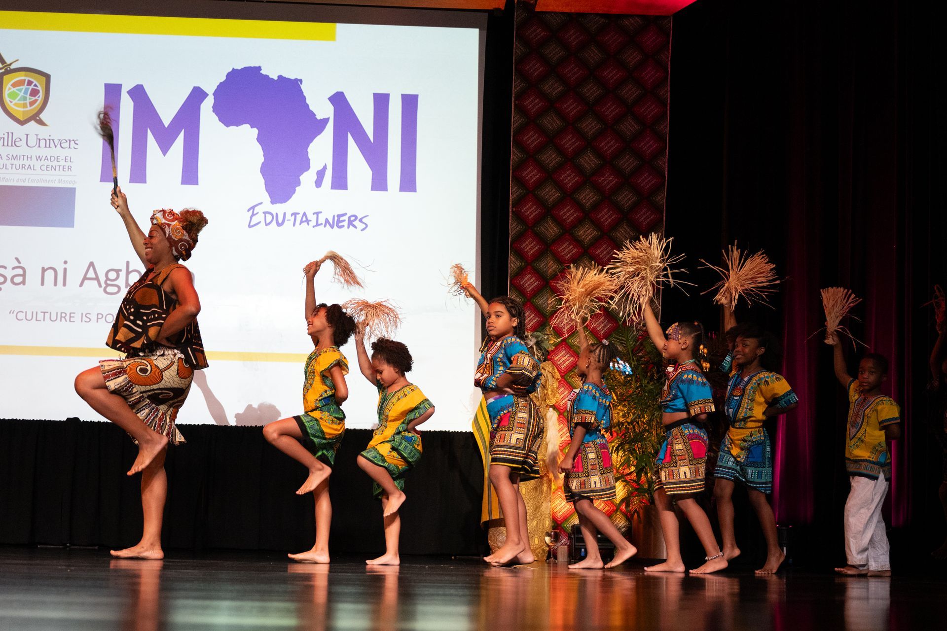 Children in colorful African clothing dance on a stage, holding fans and tall stalks, in front of a projection.