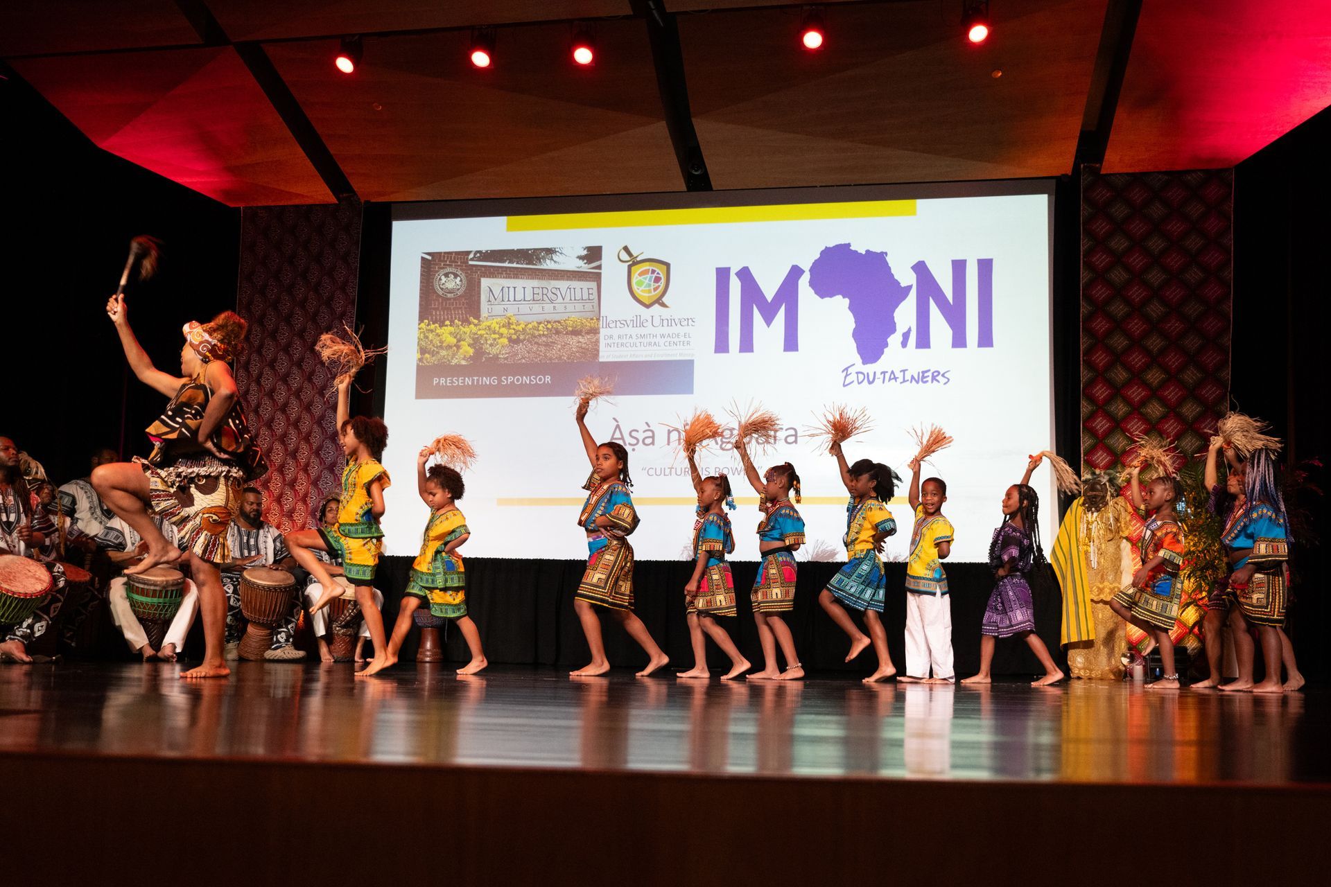 Children in colorful African attire perform a dance on a stage with a backdrop.