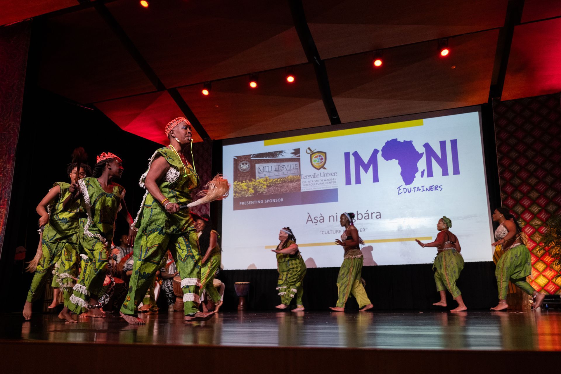 African dancers on stage, performing in colorful green outfits. A screen shows an event logo.