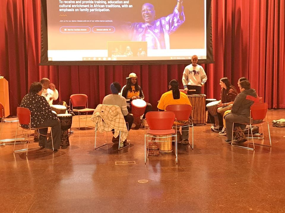 People in a circle playing drums with an instructor; presentation screen in background.
