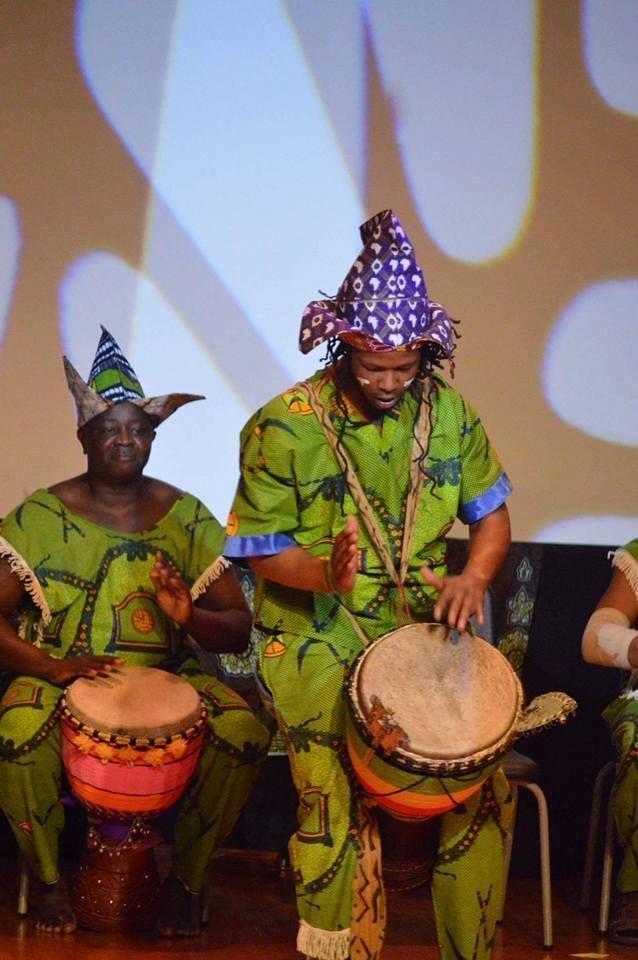 Two people in green patterned outfits play drums onstage; they wear colorful hats.