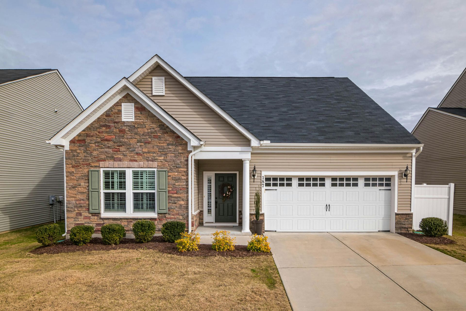 A single-story suburban house with stone and tan siding, a dark roof, a white garage, and a front porch.