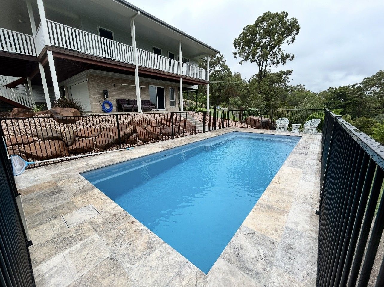 Pool in a backyard with a two-story house, stone patio, and black fence. Overcast sky.