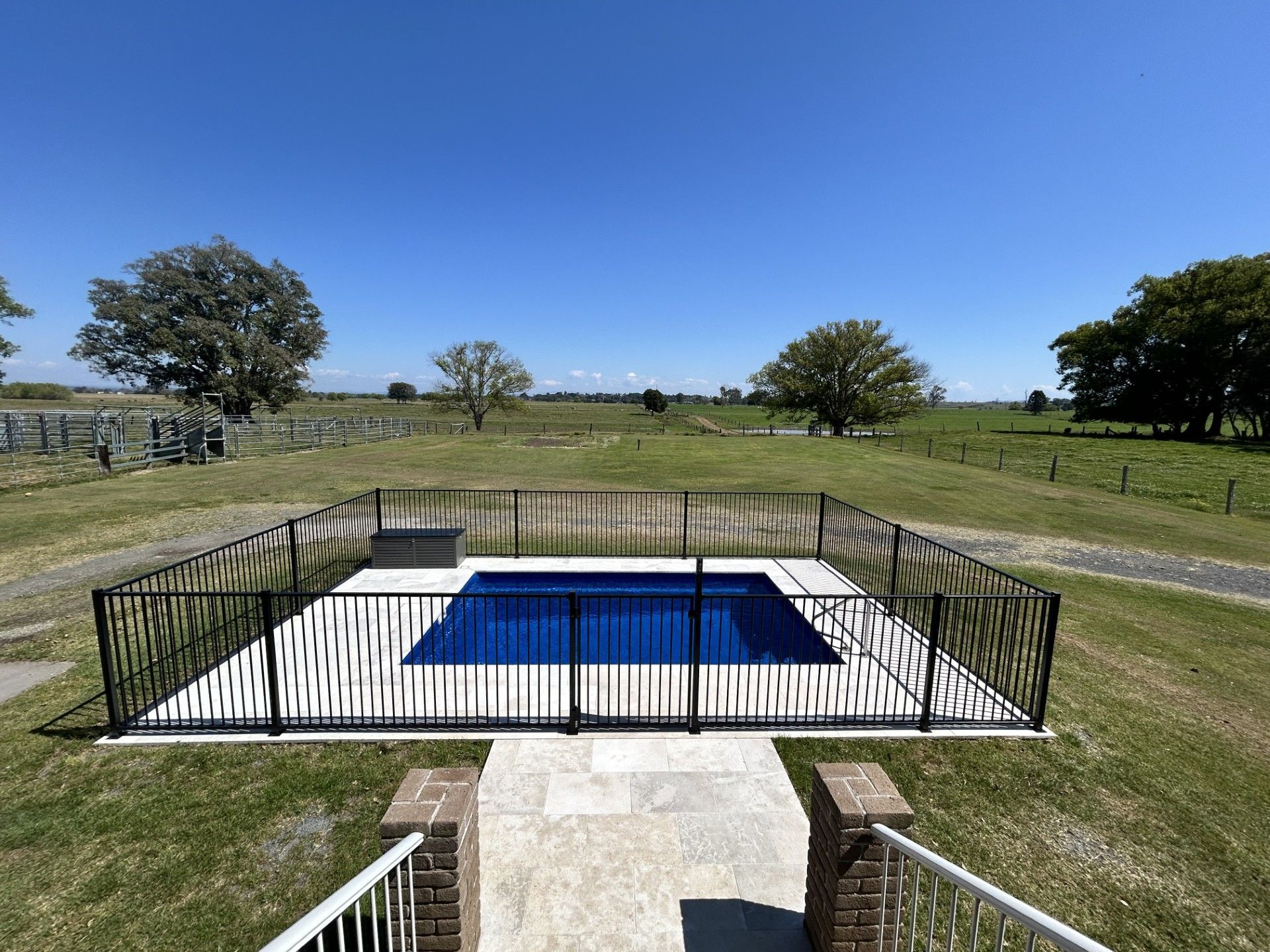 A rectangular swimming pool surrounded by a black fence on a grassy field under a blue sky.