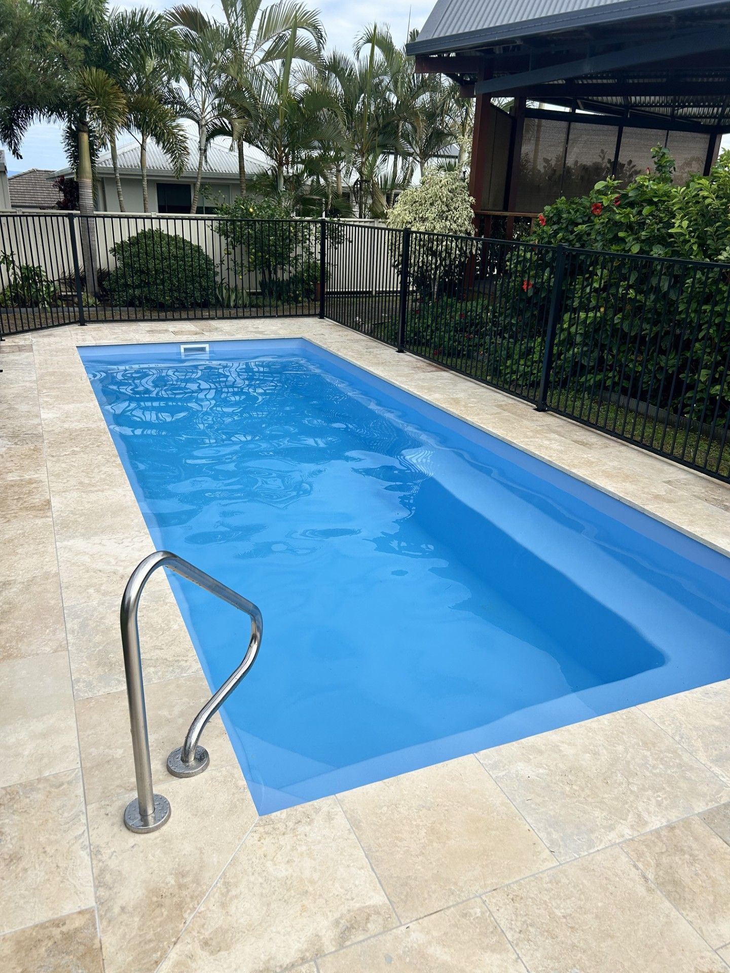Rectangular blue pool with stainless steel handrail, surrounded by stone tiles and black fence.