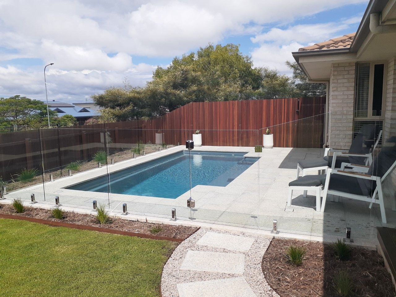 Swimming pool with glass fence, wood fence, patio, and lawn on a sunny day — Clarence Valley Pools In Yamba, NSW