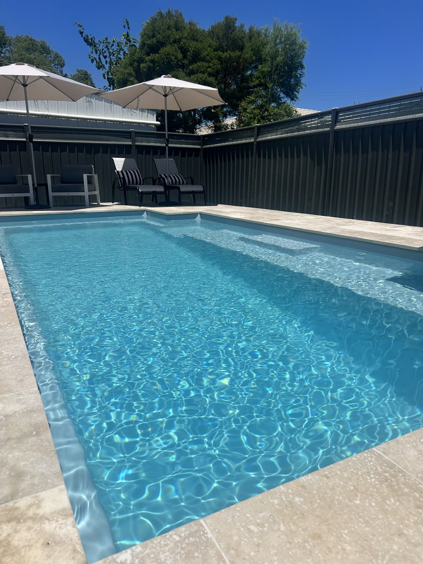A rectangular swimming pool with clear blue water and beige stone surrounds, flanked by lounge chairs under umbrellas — Clarence Valley Pools In Yamba, NSW
