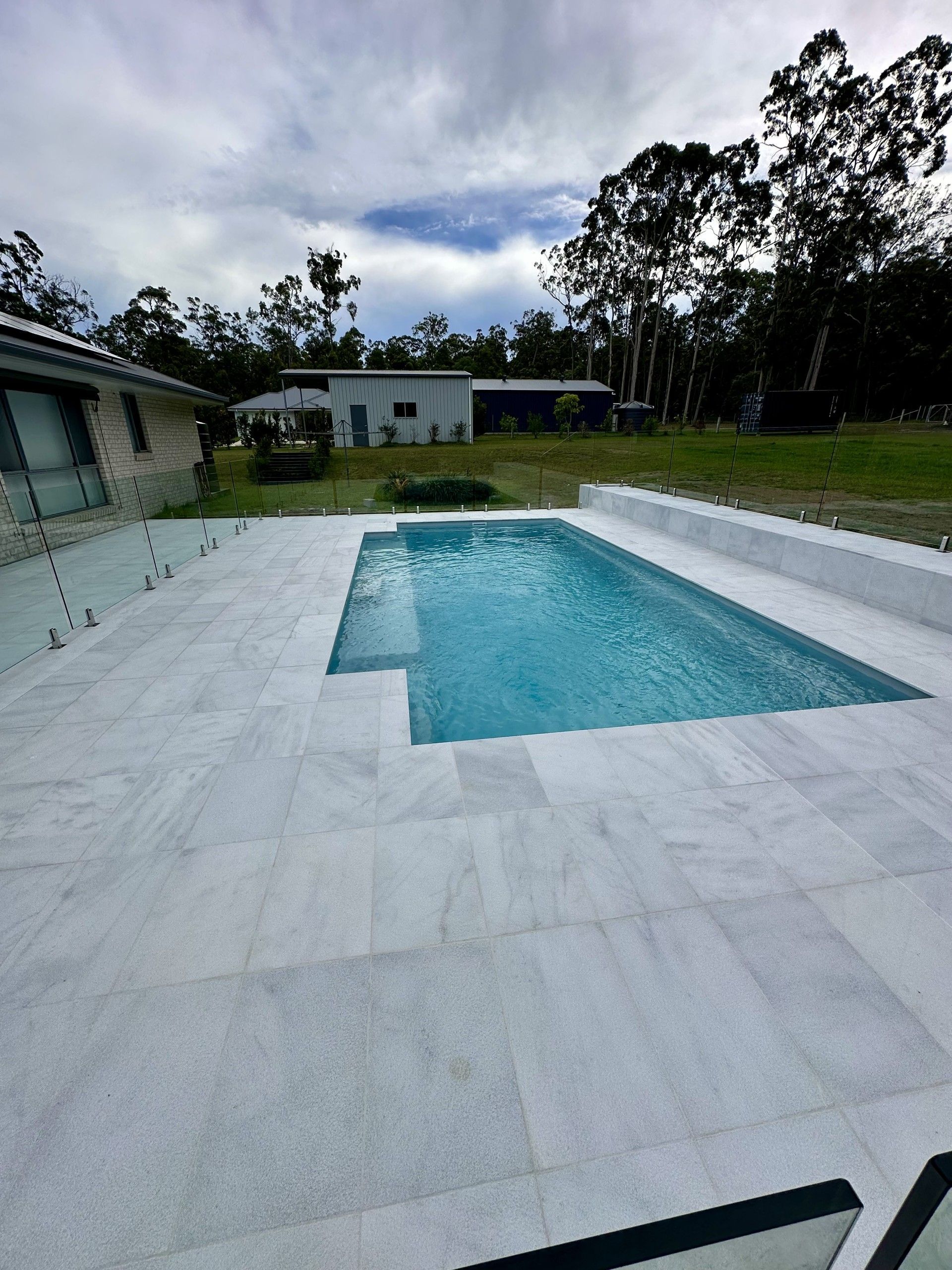 Rectangular swimming pool surrounded by light-colored stone, with a small house and trees in the background. Overcast sky — Clarence Valley Pools In Yamba, NSW