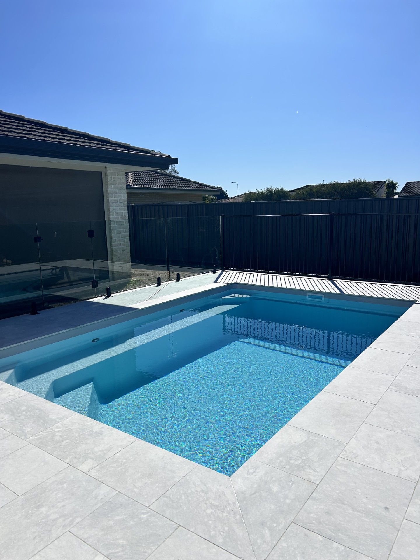 A rectangular swimming pool with blue water and light stone surround. A fence and house are in the background.