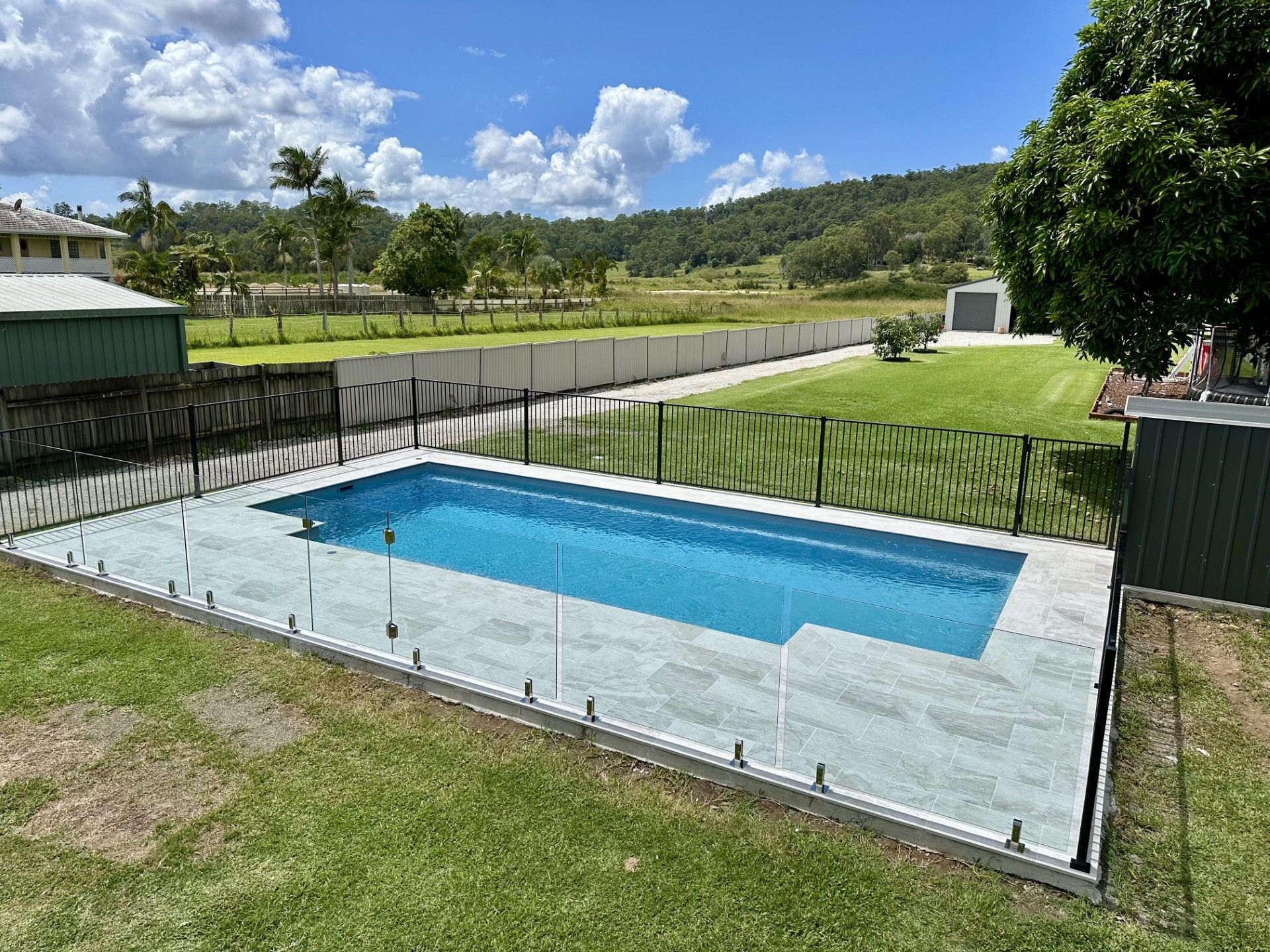 Rectangular pool with blue water and gray tile surround, enclosed by a black fence on a grassy lawn with a distant hill.