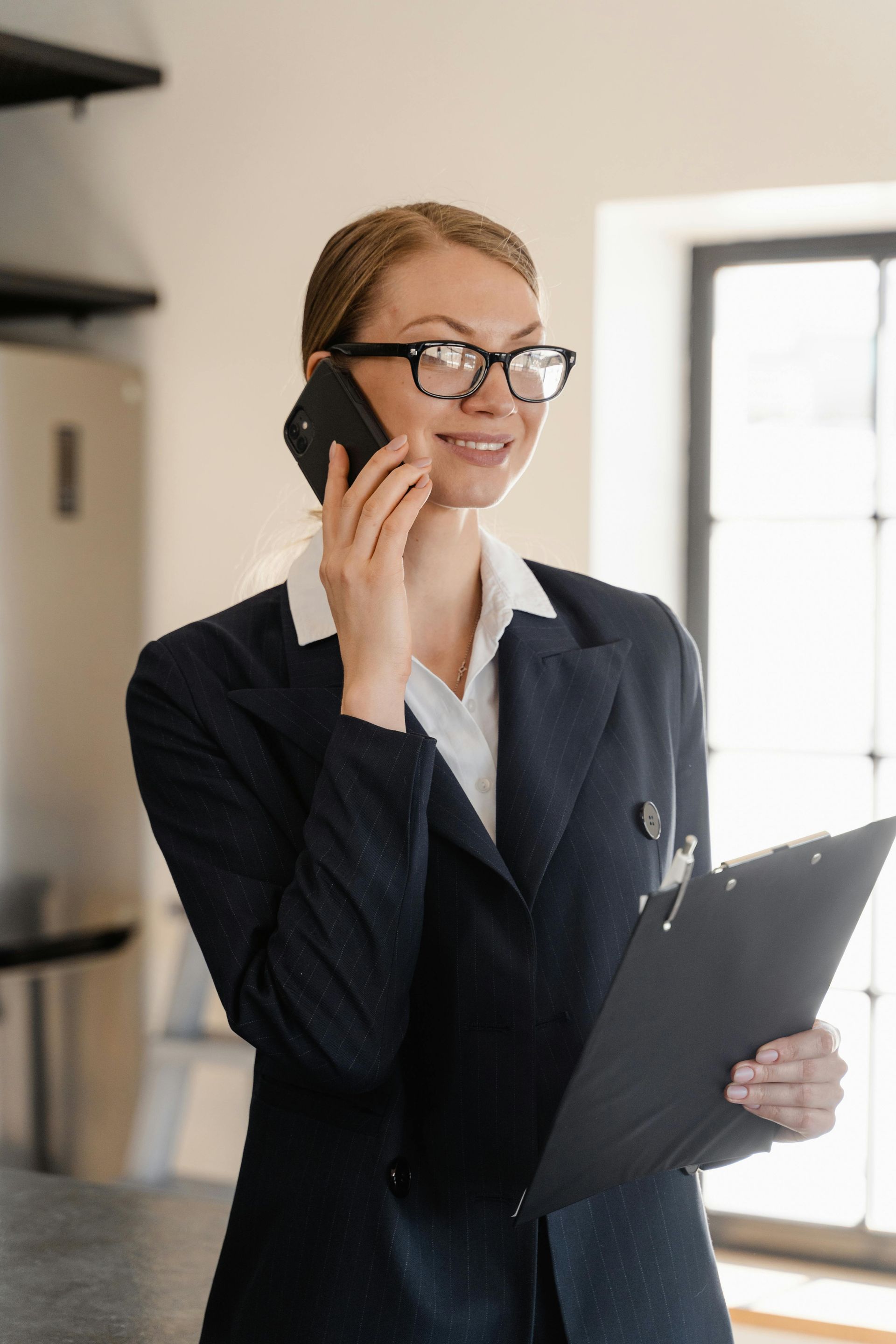 A woman in a suit is talking on a cell phone while holding a clipboard.