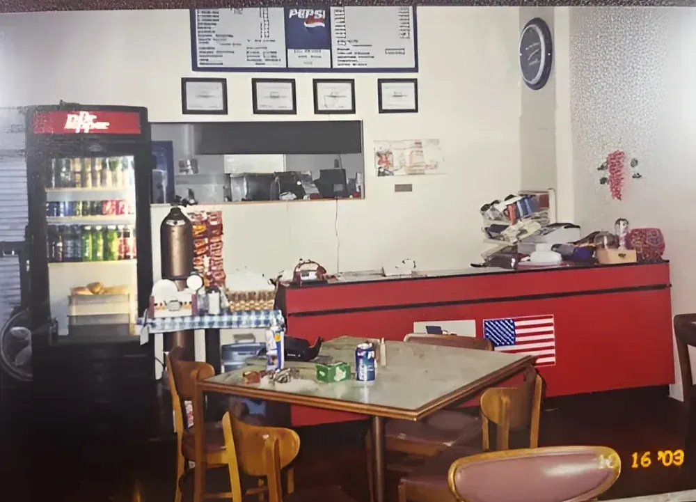 Interior of E-Z Riders from the past with a red counter, menu board, drink fridge, tables, and an American flag.