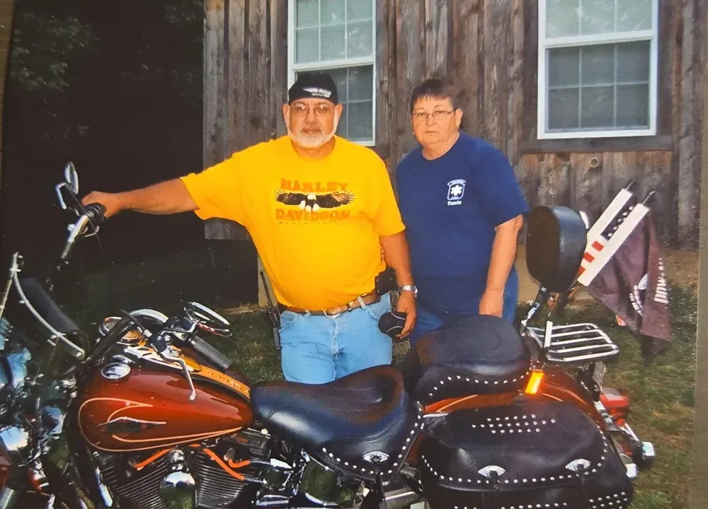 Picture of the owners with a red motorcycle in front of a wooden building.
