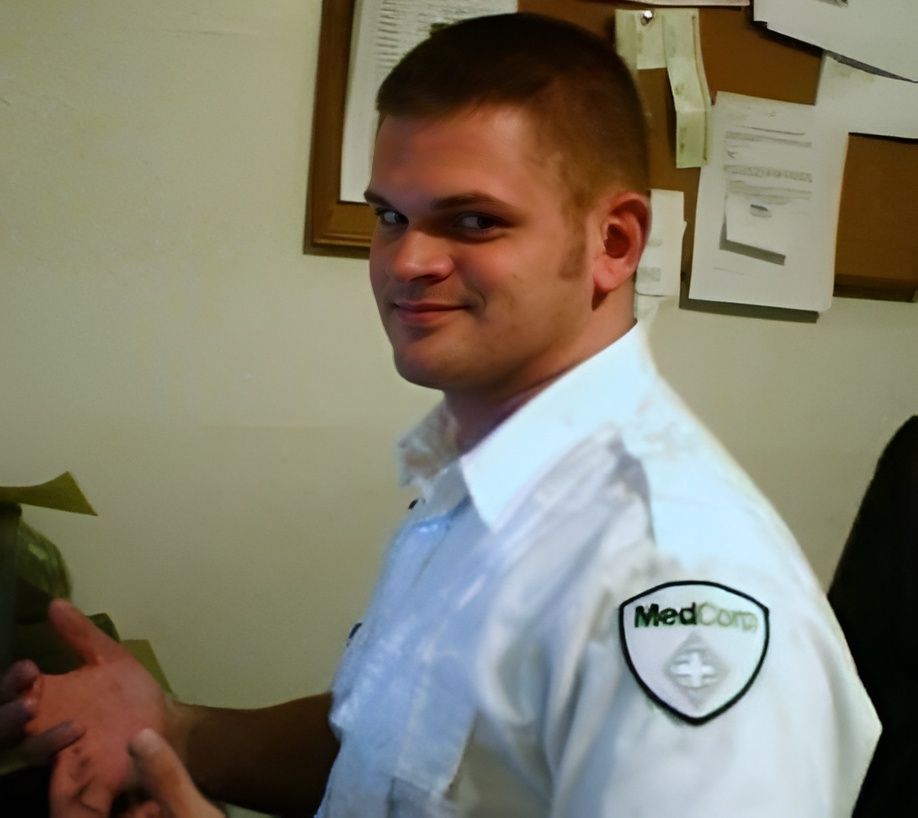 Travis in white uniform with MedCorp patch gestures, smiles in office.