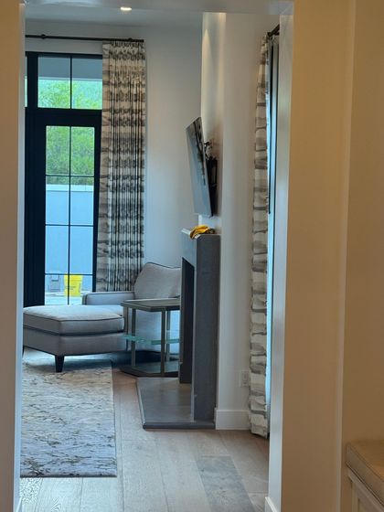 A view through a doorway into a sunlit room with a gray armchair, a fireplace, and patterned curtains flanking a window.