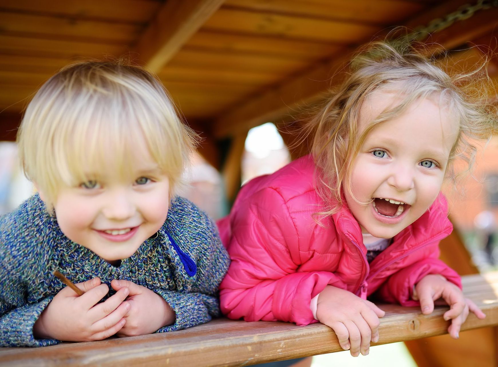 Two young children smiling, lying on a wooden surface; one wearing a blue sweater, the other a pink jacket.