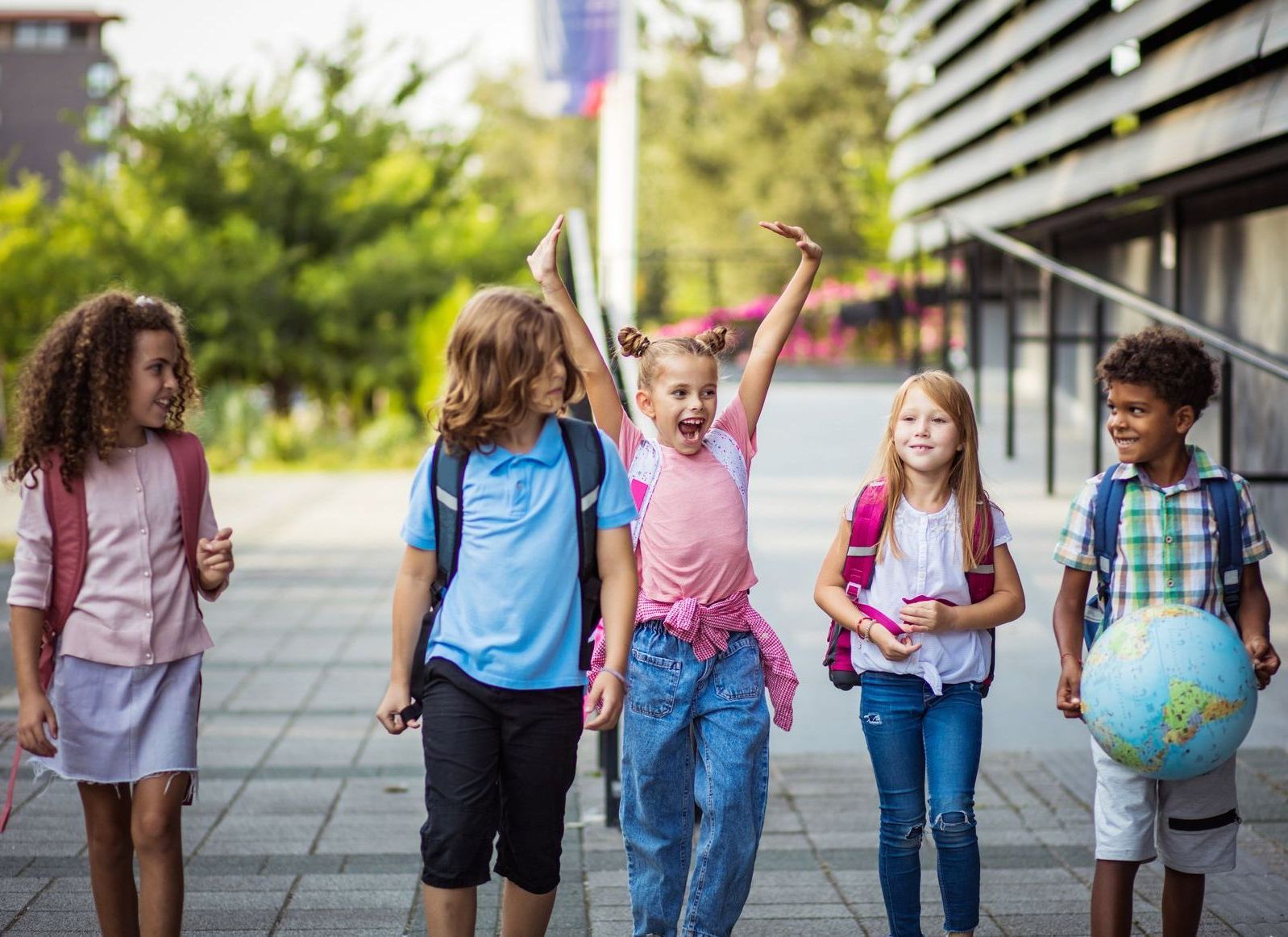 Group of children walking outside school, one raising arms in excitement, others smiling.