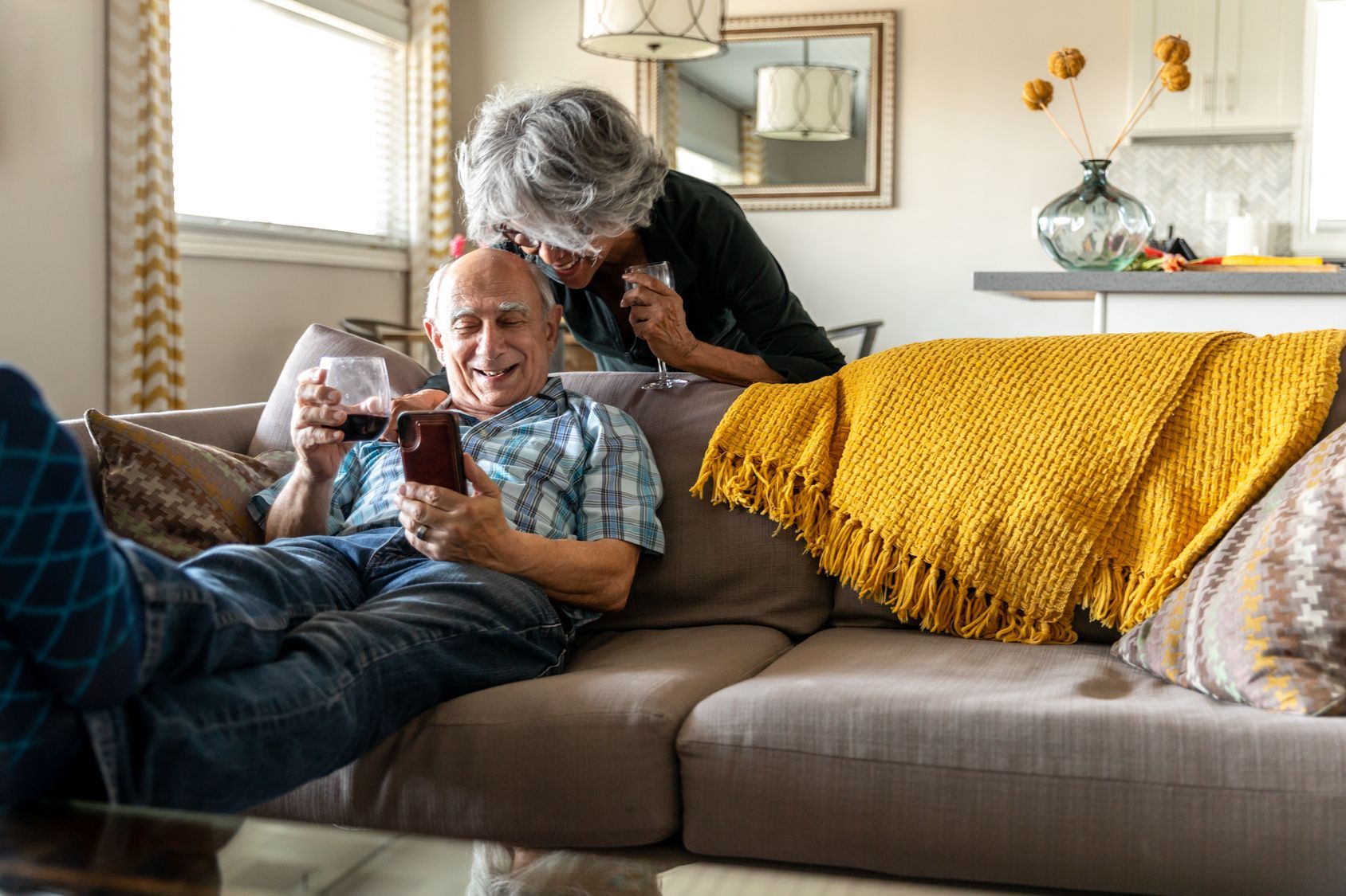 An elderly couple is sitting on a couch drinking wine and looking at a cell phone.