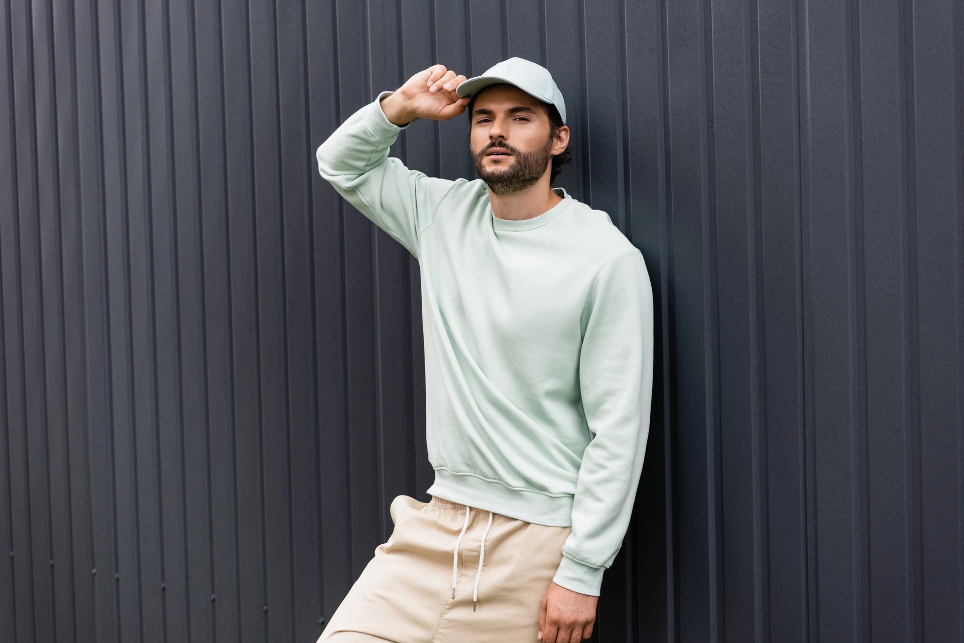 Un uomo con una felpa verde chiaro e un berretto è appoggiato a un muro scuro, con la mano sul berretto.