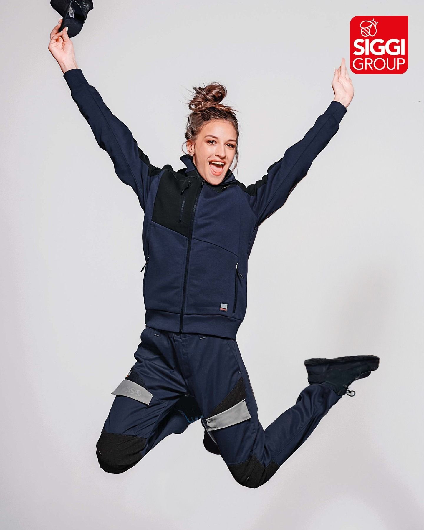 Una donna in divisa da lavoro blu salta con le braccia alzate, tenendo in mano un berretto con il logo del Gruppo Siggi.