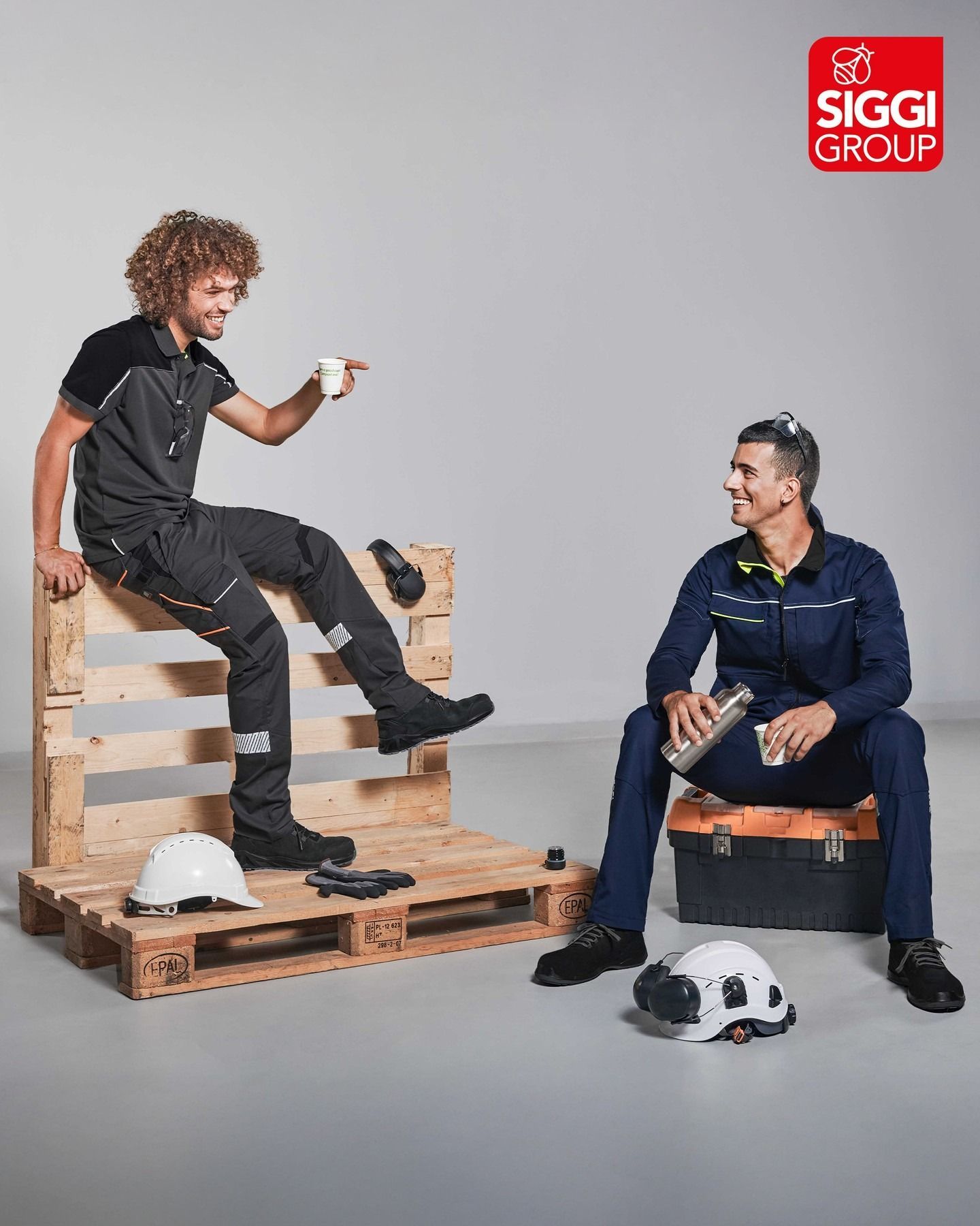 Due persone in abiti da lavoro si prendono una pausa. Una è seduta su un pallet e beve da una tazza, l'altra è seduta vicino a una cassetta degli attrezzi.