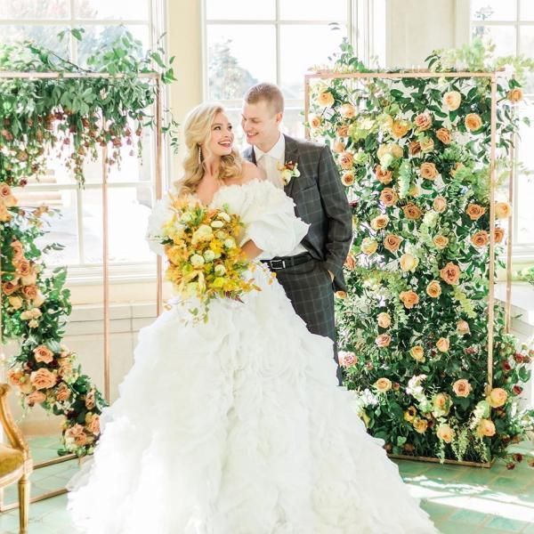 Bride and groom pose in a bright floral wedding venue, holding a bouquet and smiling.
