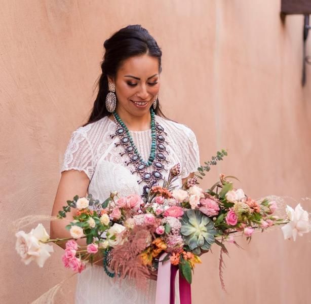 Woman holding a large pastel floral bouquet, wearing a white dress and smiling against a peach wall
