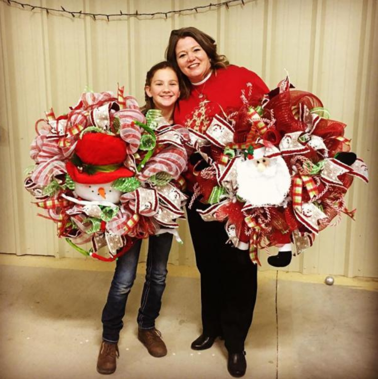 Two people standing indoors, smiling, holding large red, white, and green ribbon wreaths